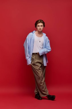 Mavi gömlekli ve pantolonlu şık bir genç adam koyu kırmızı arka planda duruyor..