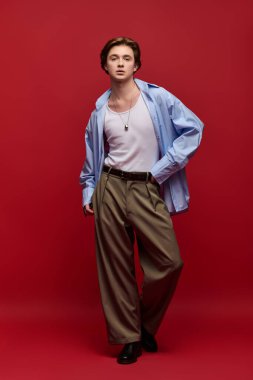 Genç adam kendine güvenerek poz veriyor. Şık bir atlet ve pantolonu kalın kırmızı arka planda..