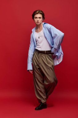 Modaya uygun genç bir adam şık bir pantolon ve koyu kırmızı bir arka planda mavi gömlekle poz verir..