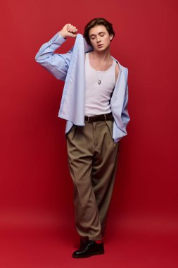 Şık bir genç adam mavi gömlekli ve pantolonlu parlak kırmızı arka planda rahatça poz veriyor..