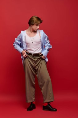 Bu genç adam şık bir mavi gömlek ve şık bir pantolon giyiyor, koyu kırmızı arka planda güvenle poz veriyor..