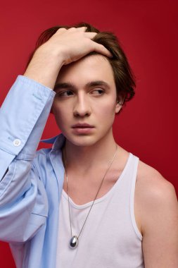 Şık bir genç adam, koyu kırmızı bir arkaplana karşı mavi gömlekli ve kolsuz bir bluzla güven içinde poz verir..