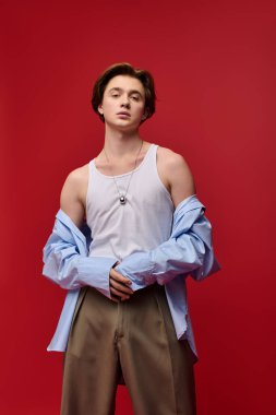 Genç adam kendine güvenerek kırmızı arka planda pantolon ve kolsuz gömlekle poz veriyor..