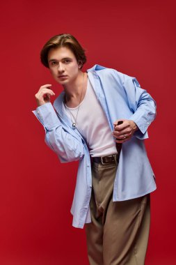 Genç bir adam şık pantolon ve mavi gömlekle poz veriyor, parlak kırmızı bir arka planı var..
