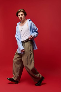 Şık genç adam kendine güvenerek poz veriyor. Moda pantolonları ve mavi gömlekli, koyu kırmızı arka planda..
