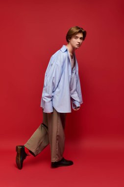 Moda bir genç adam kendine güvenerek poz veriyor. Şık pantolonlar ve mavi gömlekle. Kırmızı bir arka plana karşı..
