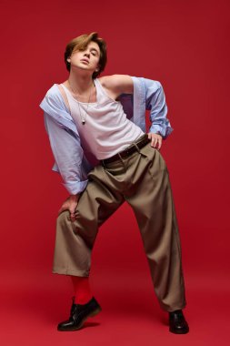 Şık, genç bir adam, kendine güvenen bir bluz ve geniş pantolonla poz veriyor, kırmızıya karşı dizilmiş..