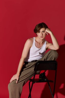 Şık bir genç adam zarif bir şekilde oturur ve şık kıyafetini kırmızı bir arka planda gösterir..