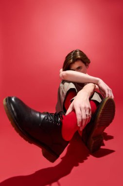 Genç bir adam pantalonla şık bir poz verir ve parlak kırmızı bir arka planda kolsuz bluz giyer..