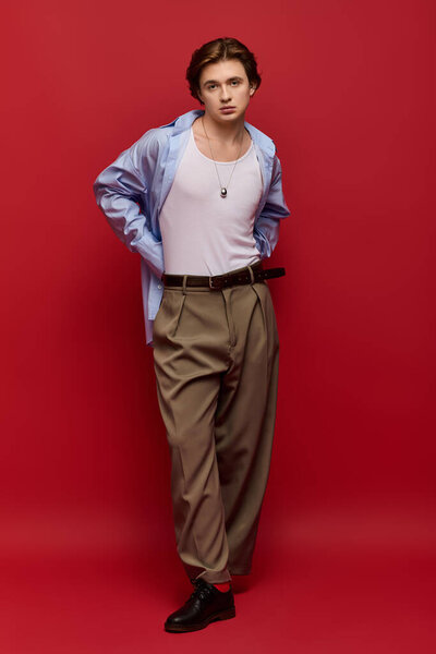 A young man poses confidently in a stylish blue shirt and pants against a red backdrop.