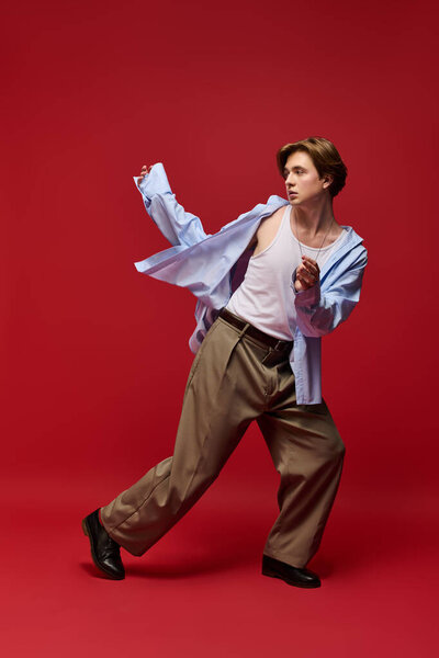 Стильный молодой человек моделирует свою синюю рубашку и свободные брюки на ярком красном фоне.