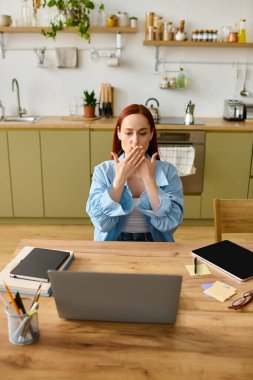 A woman with red hair teaches language online from her home kitchen, focused on her laptop.