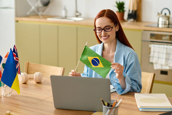 A red-haired woman teaches language online, smiling as she holds a flag while using her laptop.