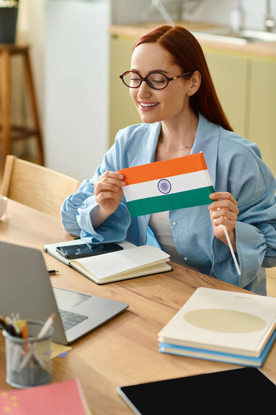 A woman with red hair teaches languages online while holding an Indian flag in her home.
