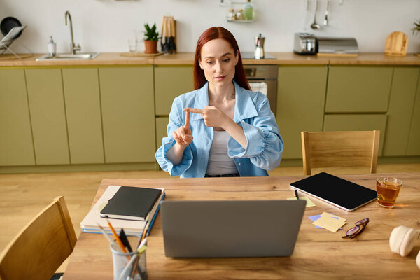 A woman with red hair teaches languages online from her kitchen, using her laptop and gestures.