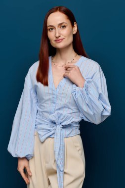 A confident redhead in a chic blouse stands out against a rich blue background.