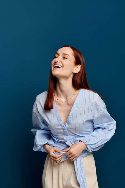 A joyful woman with red hair in a stylish blouse stands against a blue backdrop.