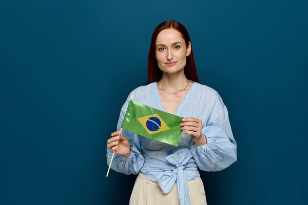 A woman with red hair holds the Brazilian flag in a bright, blue studio. She smiles confidently.