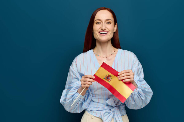 A smiling woman with red hair showcases a flag while teaching in a blue studio setting.
