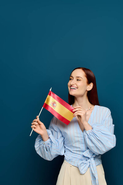 A cheerful language teacher with red hair proudly displays a flag in a blue studio environment.