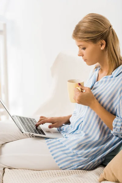 Vue latérale sur une femme adulte tenant une tasse de thé et utilisant un ordinateur portable — Photo de stock