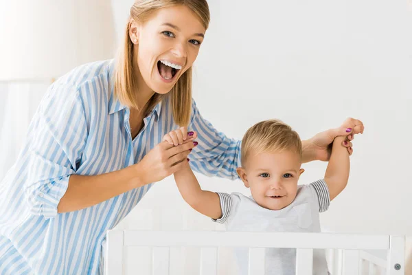 Cheerful mother looking at camera and holding smiling toddler in hands — Stock Photo