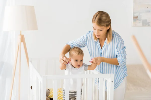 Mom holding hands of son in crib in nursery room — Stock Photo