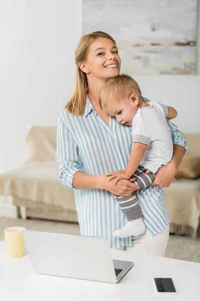 Fröhliche Mutter, die mit Laptop und Smartphone am Schreibtisch steht und lächelndes Kleinkind in den Händen hält — Stockfoto