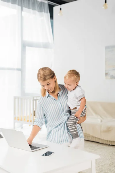 Allegra madre che tiene in mano il bambino sorridente e utilizza il computer portatile — Foto stock