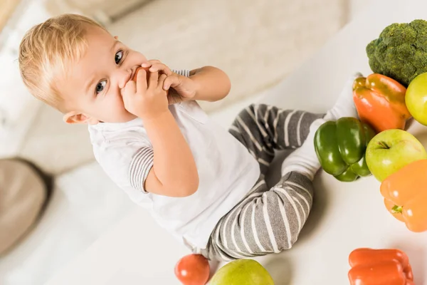 Bonito criança comendo e sentado na mesa cercada por frutas e legumes — Fotografia de Stock