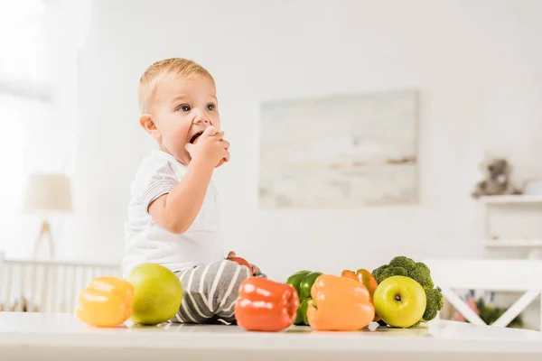 Милий малюк їсть і сидить на столі в оточенні фруктів і овочів — стокове фото