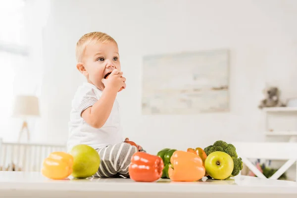 Cute toddler eating and sitting on table surrounded by fruit and vegetables — Stock Photo