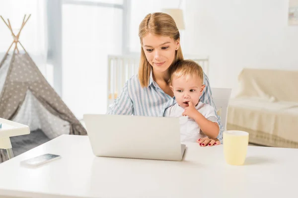Mutter sitzt mit süßem Kleinkind am Schreibtisch und benutzt Laptop — Stockfoto