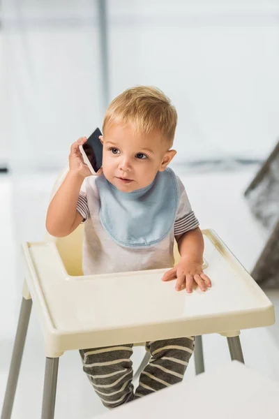 Niedliches Kleinkind spricht auf Smartphone und sitzt im Kinderstuhl — Stockfoto