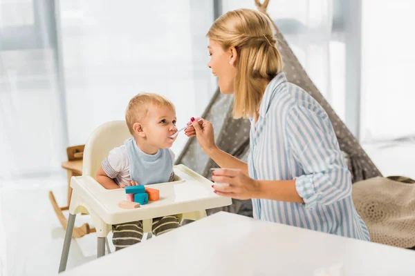 Мама кормит сына в высокий детский стульчик с детским питанием — стоковое фото