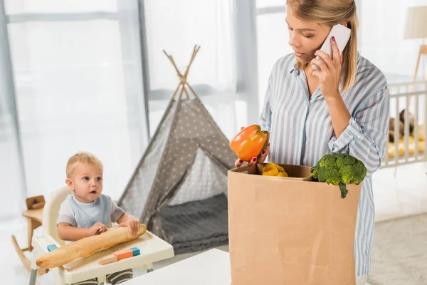 Busy mother holding groceries while talking on smartphone with toddler in baby chair on background — Stock Photo