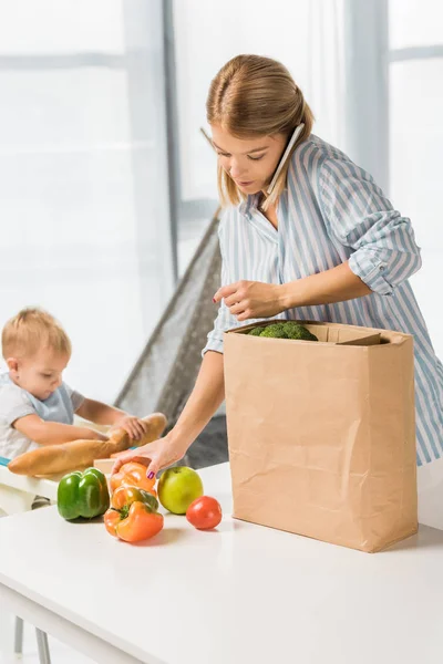 Mom holding groceries while talking on smartphone with toddler in baby chair on background — Stock Photo