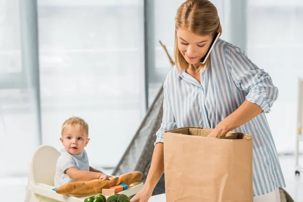 Madre sosteniendo comestibles mientras habla en un teléfono inteligente con un niño pequeño en silla de bebé en el fondo - foto de stock