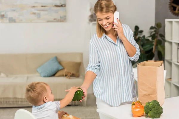 Щаслива мати тримає продукти під час розмови на смартфоні з малюком у дитячому кріслі на фоні — стокове фото