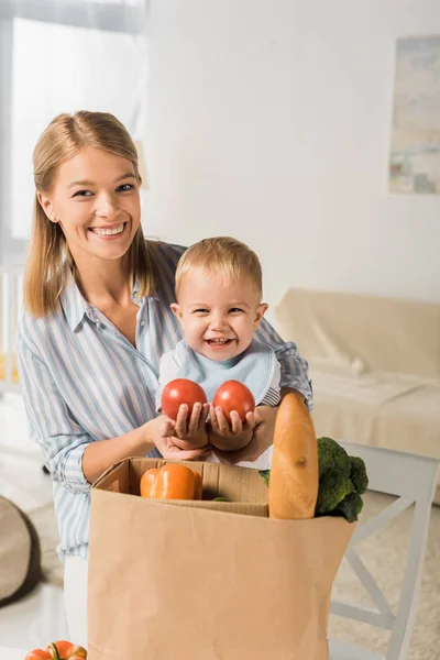 Glückliche Mutter zeigt ihrem fröhlichen Sohn Lebensmittel und schaut in die Kamera — Stockfoto