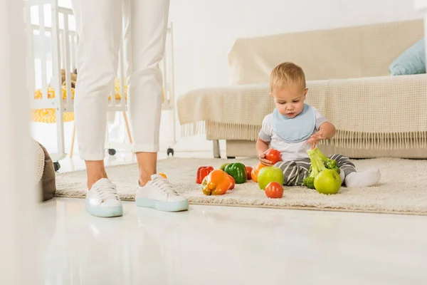 Обрезанные ноги стоят рядом с малышом, сидя на полу и играя с фруктами и овощами — стоковое фото