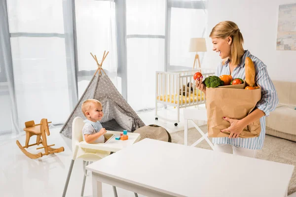 Mutter trägt Lebensmittel und schaut Sohn im Kinderstuhl an — Stockfoto