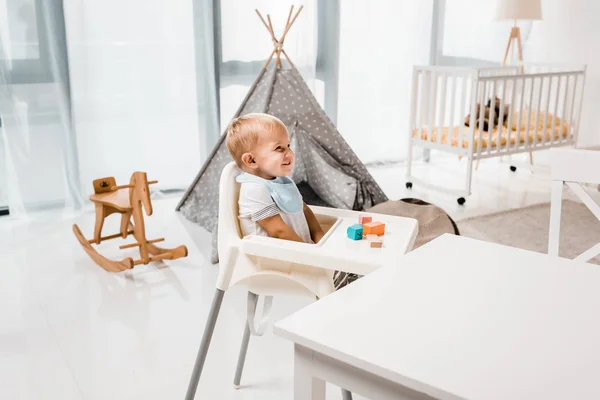 Heureux tout-petit assis dans la chaise bébé dans la chambre de pépinière avec des cubes de jouets — Photo de stock