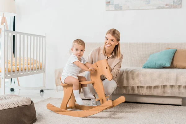 Adorable niño sentado en juguete caballo de madera y mirando a la cámara con sonriente madre - foto de stock