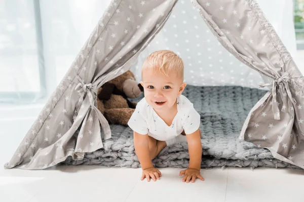 Smiling adorable toddler sitting in baby wigwam with fluffy teddy bear toy — Stock Photo