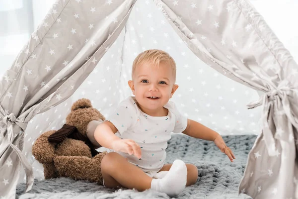 Smiling adorable toddler sitting in baby wigwam with fluffy teddy bear toy — Stock Photo