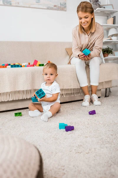 Прелестный малыш смотрит в камеру и играет с красочными кубиками и матерью — стоковое фото