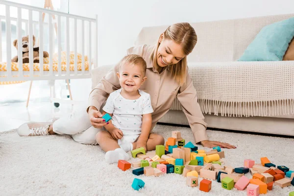 Adorable toddler playing with colorful cubes and mother in nursery room — Stock Photo