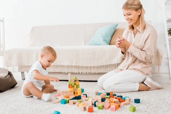 Adorable niño jugando con cubos de colores y la madre en la habitación de la guardería - foto de stock