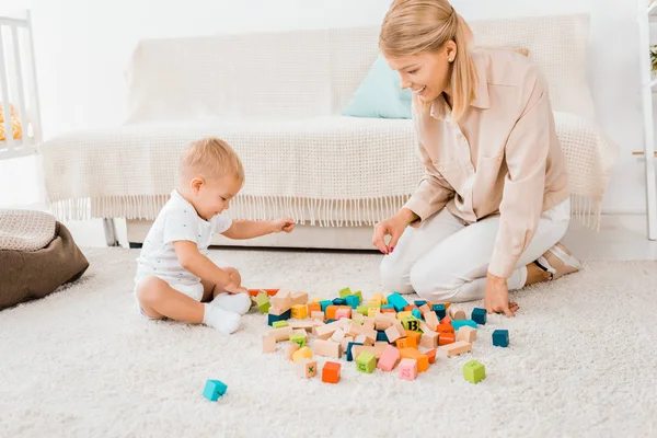 Adorable niño jugando con cubos de colores y la madre en la habitación de la guardería - foto de stock